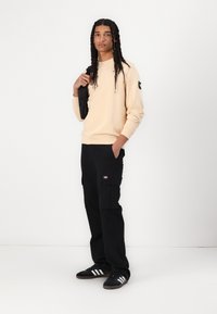 Light beige sweatshirt with ribbed cuffs, black cargo pants with multiple pockets, and black sneakers featuring white stripes.