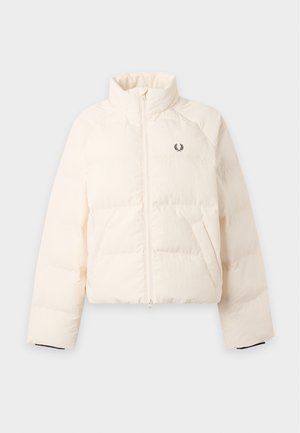 Femme portant une doudoune crème, une chemise blanche, une jupe marron foncé avec des poches avant, des chaussettes blanches et des mocassins marron foncé, debout devant un fond uni.