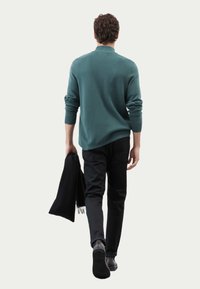 Zielony dzianinowy sweter z kołnierzem, czarne spodnie i czarny szalik w ręku. Model zwrócony plecami, ukazujący luźny krój i strukturę.