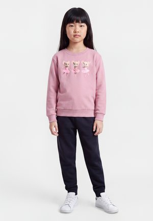 Fille portant un sweat-shirt rose avec trois illustrations de souris dansantes, pantalon noir et baskets blanches, debout sur un fond blanc.