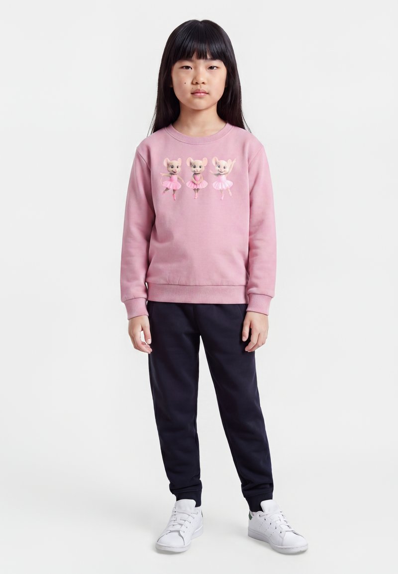 Fille portant un sweat-shirt rose avec trois illustrations de souris dansantes, pantalon noir et baskets blanches, debout sur un fond blanc.