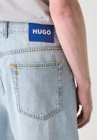 Jean en denim bleu clair avec une poche arrière visible et une étiquette HUGO bleue. Le tissu présente une texture légèrement usée et des coutures jaunes.
