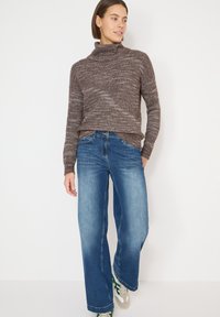 Jersey de cuello vuelto marrón de punto con un patrón texturizado, combinado con vaqueros de pierna ancha azul. El conjunto incluye zapatillas deportivas verdes y beige.