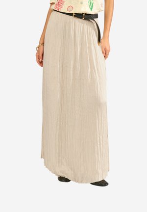 Jupe longue plissée crème avec ceinture noire portée par une personne debout sur un fond blanc, assortie d’un haut à motifs.