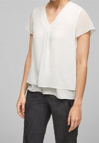 Blouse blanche légère avec un col en V, des manches courtes et un design superposé. Tissu lisse avec des détails plissés au niveau de l'encolure.