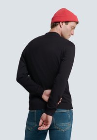 Schwarzer Strickpullover mit Rundhalsausschnitt, kombiniert mit blauen Jeans. Eine rote Mütze auf dem Kopf setzt einen Farbakzent.