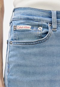 Ανοιχτό μπλε τζιν παντελόνι με κοντινή λήψη της ζώνης, η οποία περιλαμβάνει την ετικέτα Calvin Klein και ασημένια αξεσουάρ.