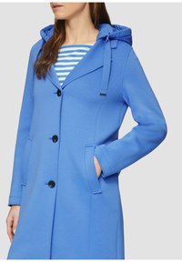 Cappotto blu con cappuccio realizzato in tessuto morbido. Presenta un colletto singolo, cinque bottoni e tasche laterali. Camicia a righe interna visibile.