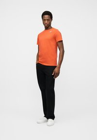 Άνδρας με πορτοκαλί κοντομάνικο πουκάμισο, μαύρο παντελόνι και λευκά αθλητικά παπούτσια, στέκεται με το ένα χέρι στην τσέπη σε λευκό φόντο.