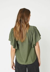 Olivgrünes Kurzarmblusenshirt mit Rüschendetails, strukturiertem Stoff und abgerundetem Saum. Kombiniert mit hochgeschnittenen dunklen Jeans, von hinten gezeigt.