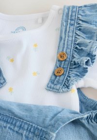 Jeanshängselbyxor med en vit ribbad topp med gula och gröna blommönster. Inkluderar två bruna knappdetaljer och samlade ärmar i denim.