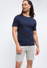 Άνδρας με μακριά dreadlocks που φοράει μπλε σκούρο κοντομάνικο μπλουζάκι και ανοιχτό γκρι σορτς, μπροστά σε απλό φωτεινό φόντο.