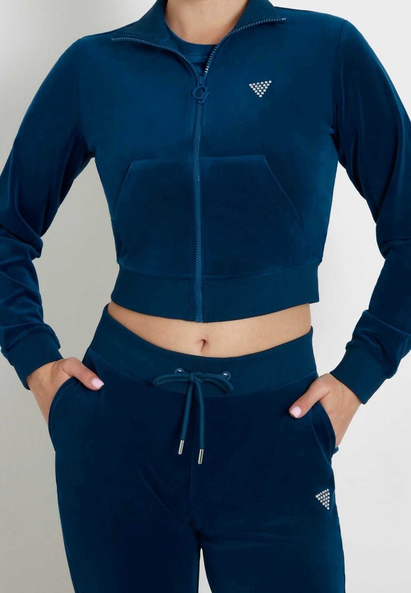 Donna che indossa una giacca cropped in velluto blu navy con zip e pantaloni abbinati con coulisse, decorati con piccoli loghi triangolari in strass, con le mani nelle tasche.