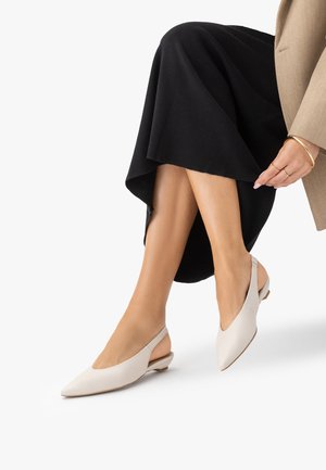 Kobieta w beżowych, szpiczastych balerinach typu slingback na niskim obcasie, skrzyżowane nogi, ubrana w czarną spódnicę i beżową marynarkę, poprawiająca rękaw marynarki.