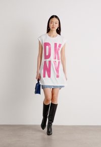 T-shirt bianco oversize con scritta bold in rosa "DKNY", gonna di jeans, stivali neri al ginocchio e una piccola borsa blu.