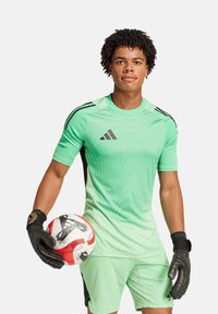 Maglia da portiere verde sportiva con design testurizzato, dettagli neri e maniche corte. Il modello tiene in mano un pallone da calcio mentre indossa guanti neri.