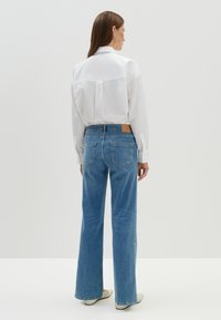 Chemise blanche à manches longues associée à un jean clair à jambes larges. Les jeans présentent une étiquette en cuir à l'arrière et deux poches arrière.