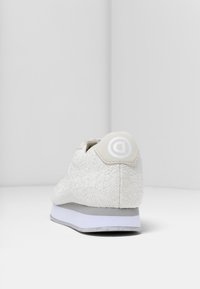 Zapatilla blanca con una superficie texturizada, que presenta un sutil patrón floral, una suela de goma y un logotipo circular en el talón.