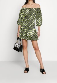 Schulterfreies Kleid mit grünen und schwarzen geometrischen Mustern, kurzen Puffärmel und einem gerafften Saum. Kombiniert mit einer schwarz-weißen Handtasche.