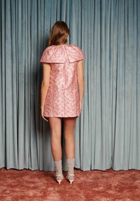 Kvinne stående med ryggen til kamera, iført en rosa kjole med blomstermønster i tekstur, gjennomsiktige sokker og hvite hæler, foran blå gardiner og brunt teppe.