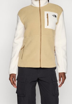 Veste en polaire beige et blanche avec fermeture éclair devant, poche poitrine zippée blanche et pantalon cargo noir avec grandes poches latérales.