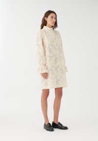 Vestido beige claro con patrón de encaje floral, cuello alto y mangas abullonadas con puños con volantes. Combinado con zapatos negros sin cordones.