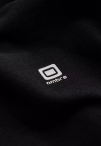 Černá tkanina s hladkým povrchem, na které je bílé logo se čtvercovým obrysem a pod ním slovo "ombre" v moderním písmu.