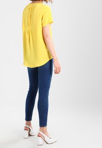 Žlutá krátká blůza s obláhem a knoflíky na zádech, spojená s tmavě modrými slim-fit džíny a bílými botami na blokovém podpatku.