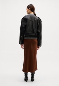 Casaco de cabedal preto de estilo motoqueiro com epauletes nos ombros, usado sobre um vestido maxi de cetim castanho, combinado com botas pretas de salto alto até ao joelho.