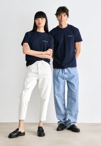 Two people standing side by side wearing navy blue "tommy jeans" T-shirts, white and light blue jeans, and black slip-on shoes against a plain background.