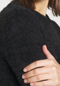 Osoba w czarnej, puszystej bluzie trzyma jedną ręką górną część ramienia, krótka brązowa fryzura widoczna w okolicach szyi.