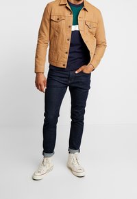 Vaaleanruskea denimtakki, jossa on kaksi rintataskua, alla laivastonsininen ja vihreä raidallinen paita, tummansiniset farkut ja valkoiset korkeavartiset tennarit.