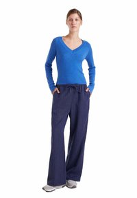 Jersey azul de punto con escote en V, combinado con pantalones de pierna ancha a rayas finas en azul oscuro. Modelo con zapatillas grises, manos en los bolsillos.