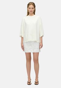 Biely oversize tričko vyrobené z jemného materiálu, spárované s bielou čipkovanou sukňou. Modelka stojí s minimálnymi doplnkami a plochými čiernymi sandálmi.