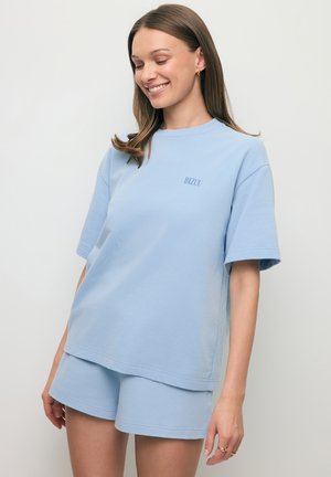 Vrouw die een lichtblauw oversized T-shirt met korte mouwen en bijpassende korte broek draagt, glimlacht met gesloten ogen tegen een effen achtergrond.