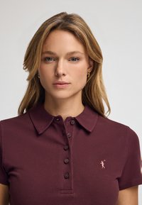 Burgundowa koszulka polo z kołnierzykiem i krótkimi rękawami, z pięcioknapowym zapięciem oraz małym różowym logo po lewej stronie klatki piersiowej.