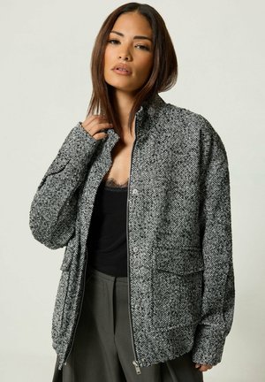 Femme portant une veste grise à motif chevron sur un haut noir et un pantalon gris, tenant le col de la veste d'une main.
