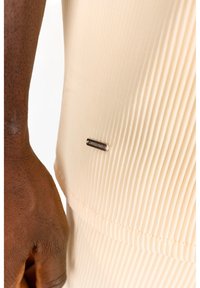Top in tessuto beige a costine con un piccolo tag logo argento. La texture presenta scanalature verticali e un design aderente. Mani parzialmente visibili.
