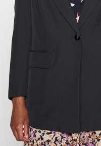 Black blazer in smooth fabric, featuring a single button closure, notch lapels, and a front pocket, paired with a floral patterned skirt.