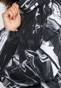 Zwart-witte jas met een glanzend, abstract patroon, voorzien van elastische manchetten en een ritssluiting aan de voorkant, gemaakt van lichtgewicht materiaal.