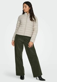 Gai bēša pufīgs jaka ar horizontālām ielām, augstu apkakli un priekšu ar rāvējslēdzēju, saskaņota ar olīvu zaļiem kārtojuma platām biksēm un melnām kurpēm ar papēdi.