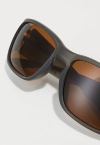 Szare okulary przeciwsłoneczne z dużymi brązowymi szkłami i matowym wykończeniem. Design charakteryzuje się grubą oprawką i prostą linią brwi.