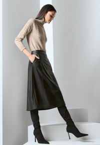 Beige Strickpullover mit hohem Kragen, kombiniert mit einem schwarzen Leder-Midikleid mit seitlichen Taschen und schwarzen Stiletto-Stiefeln über dem Knie.