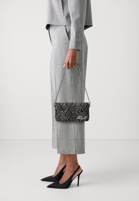 Musta-valkoinen käärmekuvioinen käsilaukku ketjuhihnalla, pidetään yhdessä kädessä. Asussa on ruutukuvioinen puku ja mustat korkokengät.