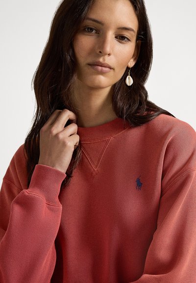 Rdeč bombažni pulover z žepi in izrezom z rebrastim robom, na prsih z majhnim modrim logotipom. Tekstura je videti mehka in sproščena.