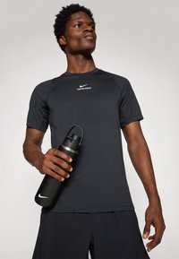 Mann i svart Nike Pro skjorte og shorts som holder en svart Nike vannflaske, ser selvsikkert oppover mot en ensfarget bakgrunn.