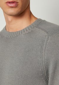 Grå stickad tröja med rund halsringning, strukturerade ribbade manschetter och axelsömmar. Har subtila stickningsdetaljer och en mjuk yta.