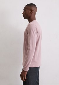 Młody mężczyzna stojący w profilu, ubrany w jasnoróżową koszulę z długim rękawem i ciemne spodnie, na tle gładkiej białej ściany.