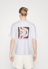 Balta medvilnė t-shirt su daugiakryptėmis grafikinėmis spaudėmis ant nugaros, su užrašu "SOUTH SURF UNION" ir kreivomis linijomis rudos, geltonos ir purpurinės spalvoms.