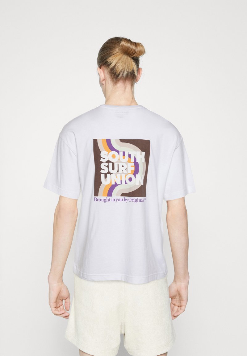 Balta kokvilnas T-krekls ar daudzkrāsainu grafiku uz muguras, kurā ir uzraksts "SOUTH SURF UNION" un izliekta līnijas brūnā, dzeltenā un purpursarkanā krāsā.